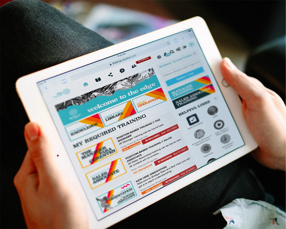 Holding ipad with screenshot of Wisetail LMS on it. Screenshot is of the website, Bridger Bike and Ski's LMS/LXP: The Edge. At the top is a banner reading "Welcome to The Edge". Then there are three modules below: Product Knowledge, Resource Library, and Shop Operations. Below this is required training, presented in English and Spanish. On the right column is a space to redeem purchased access codes, a button for Manager Training, a button for Sales Associate training, and quick links for employees.