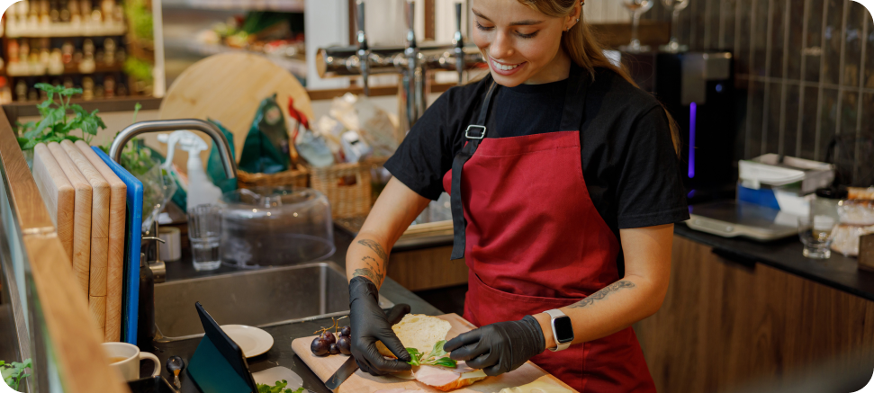 A woman in a red apron is preparing a sandwich in a cafe, wearing gloves and smiling while placing ingredients on bread, with a mobile tablet on the table in front of her.